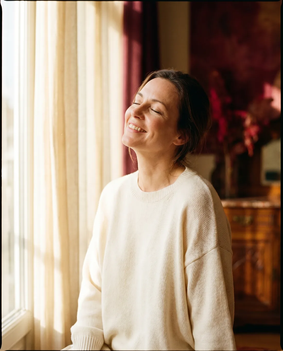 A woman resting in warm window light, eyes closed and at ease, the feeling of being well